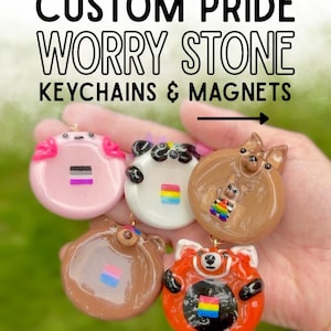 May include: Five round, glossy, custom pride worry stone keychains and magnets featuring sculpted animal faces including an axolotl, panda, dog, bear, and red panda. Each piece displays a small pride flag. Text reads: CUSTOM PRIDE WORRY STONE KEYCHAINS & MAGNETS.