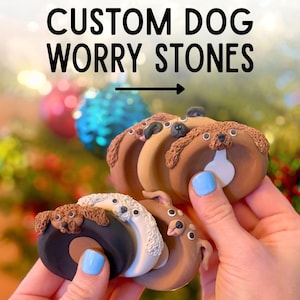 May include: A collection of custom dog worry stones in various colors and designs. The stones are donut-shaped with a dog's face on top. The text "CUSTOM DOG WORRY STONES" is displayed above the stones.
