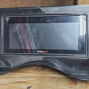May include: Black dashboard housing with a rectangular screen. The screen displays the Holley EFI logo. The housing has a textured surface and a curved bottom edge. Likely for a vehicle.