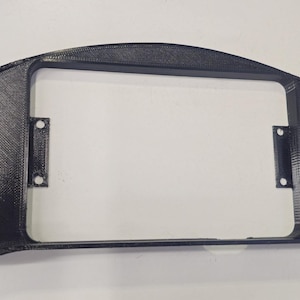 May include: A black, 3D-printed dashboard bezel with a rectangular display aperture. The bezel has a textured surface and mounting holes. The curved shape has a symmetrical design. The dimensions are in centimetres.
