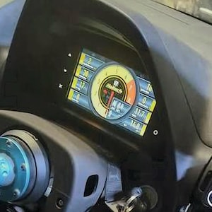 May include: A digital vehicle dashboard display featuring a prominent yellow circular tachometer with a red needle, surrounded by various numerical readouts and status indicators, set within a dark gray housing behind a steering column hub.
