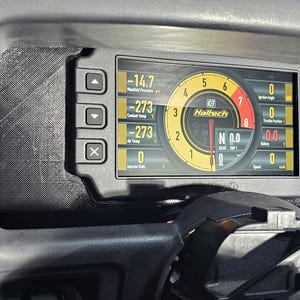 May include: Dashboard display screen with digital gauges and the brand name "Haltech". The screen shows various readings, including temperature and pressure, with a circular gauge in the center. The display is set in a black dashboard.