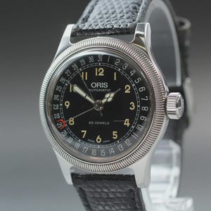 Puede incluir: Un reloj de pulsera automático Oris con esfera negra, manecillas plateadas y números arábigos. El reloj cuenta con una manecilla de fecha con punta de media luna roja, un bisel plateado estriado y una correa de cuero negro texturizado.