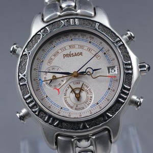 May include: Silver-toned Presage watch with a light-colored face. The watch features multiple subdials, including a calendar and alarm function. The bezel has Roman numerals and a textured design. The watch band is also silver-toned.