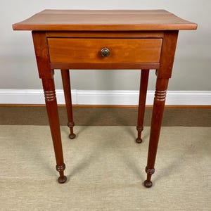 May include: A small, antique wooden side table with a single drawer and four slender, turned legs. The table is a warm reddish-brown color, with a brass knob on the drawer. The legs have decorative detailing near the top and bottom.