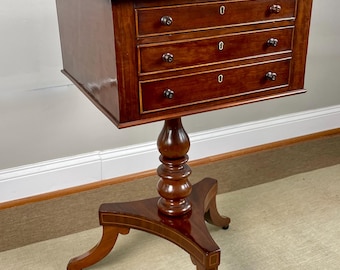 May include: A mahogany side table with three drawers and a pedestal base. The table has a rectangular top and a decorative inlaid design on the legs. The drawers have small, round knobs. The table is approximately 30 inches tall.