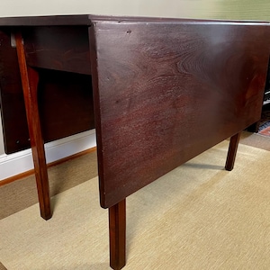 May include: A dark brown wooden drop-leaf table featuring a rectangular top with two hinged side panels that fold downward. The table is supported by four straight legs, with two legs positioned to swing outward to provide structural support.
