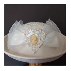 Op de afbeelding: Een witte vilt hoed met witte kant en parelversiering. Een witte satijnen strik met een cameo is gecentreerd op de hoed.