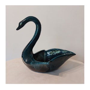 Puede incluir: Un macetero de cerámica en forma de cisne azul turquesa con un acabado brillante. El cuello del cisne está curvado y el macetero tiene un cuenco ancho y poco profundo.