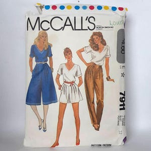 Può includere: Un pacchetto di modelli di cucito McCall's vintage con illustrazioni di modelli di abbigliamento femminile. Il pacchetto mostra una tuta blu, un completo pantaloncini bianco e pantaloni marroni con un top bianco. Il numero del modello è 7911.