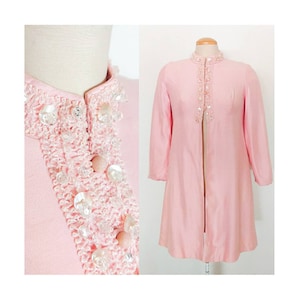 Op de afbeelding: Een roze vintage jas met een kraag van kralen en een lange, open voorkant. De jas is gemaakt van een zijdeachtige stof en heeft een klassieke, elegante stijl.
