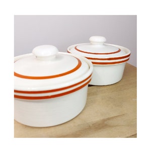 Puede incluir: Dos cuencos de cerámica blanca con rayas naranjas y tapas. Los cuencos están sobre una superficie de madera.