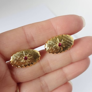 May include: A pair of gold oval cufflinks with a red rhinestone in the centre of each. The cufflinks have a textured design.
