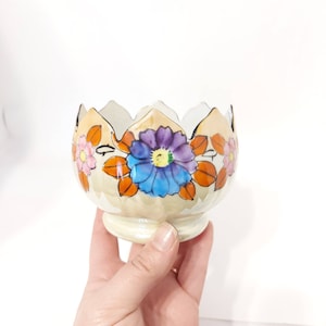 May include: A small, cream-coloured ceramic bowl with a scalloped rim. The bowl is decorated with hand-painted flowers in shades of pink, blue, purple, and orange.
