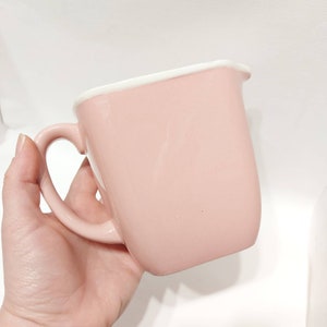 Puede incluir: Una jarra de crema de cerámica rosa con un borde blanco y un asa. La jarra está vacía y lista para llenarse con leche o crema.