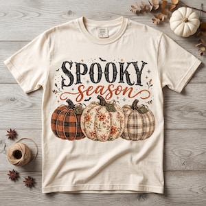 Peut inclure: Un t-shirt à manches courtes couleur crème avec le texte noir et orange "SPOOKY season" au-dessus de trois citrouilles. Les citrouilles présentent des motifs différents : un carreau noir et orange, un imprimé floral et un motif écossais beige.