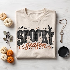 May include: A cream-coloured t-shirt featuring the text "SPOOKY season" in a black and grey buffalo plaid pattern. The design includes illustrations of two white ghosts, a black cat, bats, a spider, and small stars in orange and black.