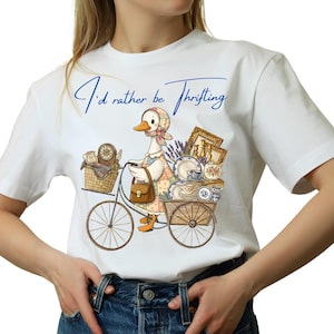 Peut inclure: T-shirt blanc avec l'inscription bleue "I'd rather be Thrifting". L'illustration représente une oie à vélo transportant des objets vintage : assiettes, cadres et un panier. Un design charmant pour les amateurs de brocante.