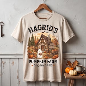 May include: A beige short-sleeve t-shirt featuring a graphic of a house, pumpkins, and a ghost holding a mug. The text reads HAGRID'S EST. 1981 PUMPKIN FARM MAGICALLY GROWN hand picked with care, with signs for PUMPKINS, HAY RIDES, HOT CIDER, APPLE PICKING.