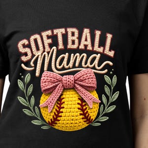 May include: Black t-shirt featuring the text "SOFTBALL Mama". The design includes a pink bow, a yellow softball with red stitching, and green leaves. The shirt is made of a soft material. The design is centered.