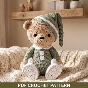 May include: A crocheted teddy bear toy featuring tan yarn for the head and limbs, wearing a sage green nightcap with a white trim, a sage green onesie with a white collar, two white buttons, and white crocheted booties. Text reads PDF CROCHET PATTERN.