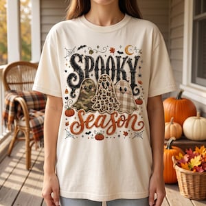 May include: A cream-coloured t-shirt featuring a graphic print with the text "SPOOKY SEASON" in black and orange. The design includes three ghosts with camouflage, leopard print, and plaid patterns, surrounded by pumpkins, bats, and spiderwebs.