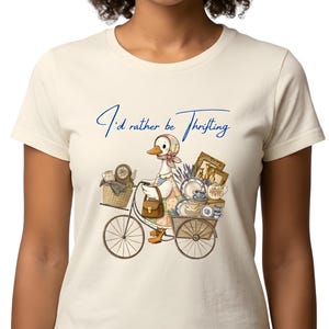 Peut inclure: T-shirt crème avec une illustration fantaisiste d'une oie à vélo, chargée de trésors vintage. Le texte "I'd rather be Thrifting" est écrit au-dessus de l'oie. Le vélo transporte un panier, un sac à main et divers objets anciens.