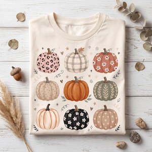 Peut inclure: Un t-shirt couleur crème arborant un imprimé graphique de neuf citrouilles aux motifs et couleurs variés, incluant léopard, carreaux, fleurs, pois et orange uni, disposées en grille avec de petites étoiles, cœurs et spirales.