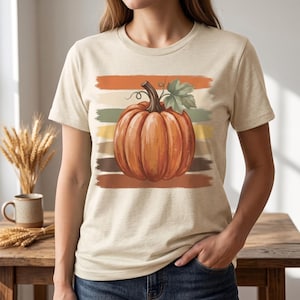 May include: A beige short-sleeve t-shirt featuring a central graphic of a large orange pumpkin with a green leaf and stem. The pumpkin is layered over horizontal brushstroke stripes in shades of orange, yellow, green, and brown.