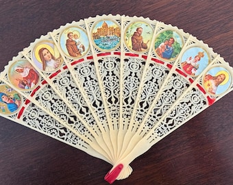 May include: Ivory-colored hand fan with intricate cut-out designs and red ribbon accents. The fan features circular illustrations of religious figures and scenes. A decorative item.