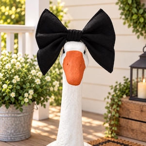 May include: A white goose statue featuring a large, solid black fabric bow accessory positioned on its head. The goose has a textured white body and a smooth, solid orange beak. The bow is oversized with pointed ends and a central knot detail.