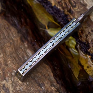 May include: A folding knife handle featuring a metallic spine with a decorative, scalloped, and notched pattern. The handle is constructed with dark, smooth side panels and a central brass-toned inlay that runs along the length of the spine.