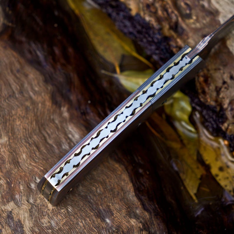 May include: A folding knife handle featuring a metallic spine with a decorative, scalloped, and notched pattern. The handle is constructed with dark, smooth side panels and a central brass-toned inlay that runs along the length of the spine.