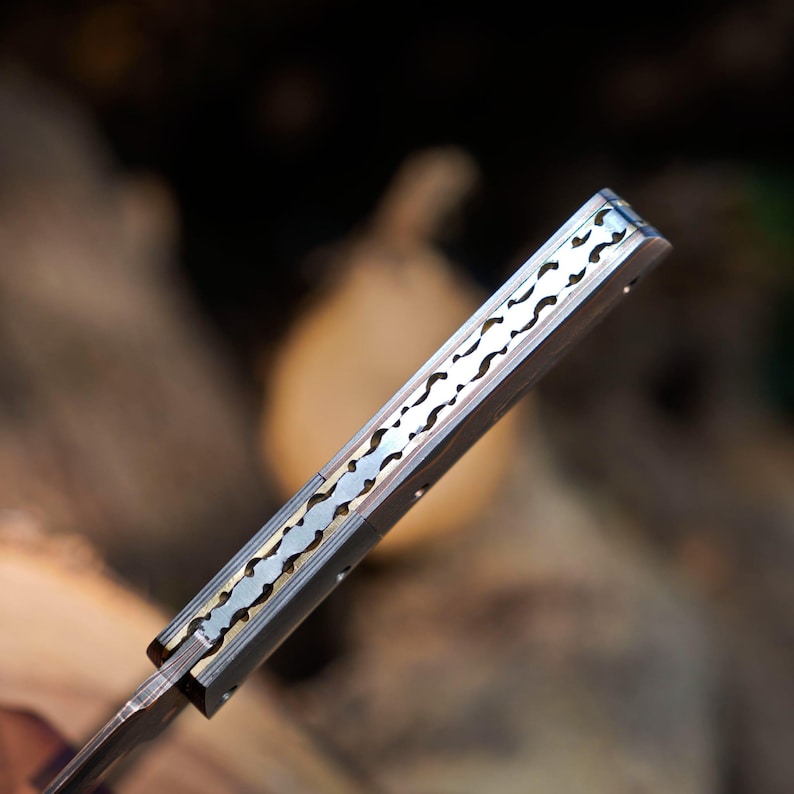 May include: A folding knife handle featuring a metallic spine with a decorative, intricate cut-out pattern. The handle is constructed from smooth, light-colored material with visible rivets, showcasing a detailed, scalloped metal inlay along the top edge.