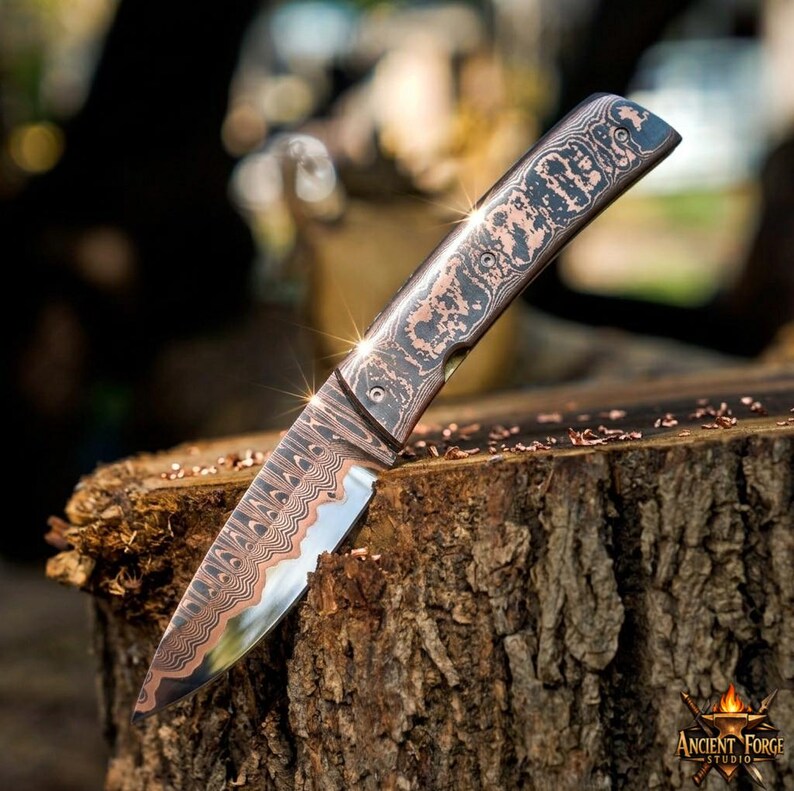 May include: A folding knife featuring a blade and handle with a distinct copper and dark metal layered pattern. The metal surface displays a wavy, intricate design throughout the entire length of the tool, showcasing a metallic finish.