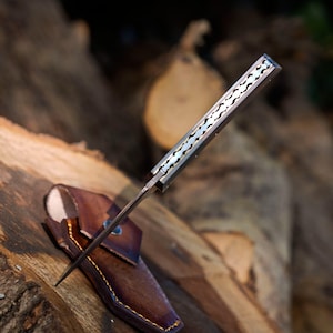 May include: A folding knife with a metallic handle featuring a decorative scalloped spine pattern. The knife is partially inserted into a brown leather sheath with visible yellow stitching and a metal rivet detail on the front flap.