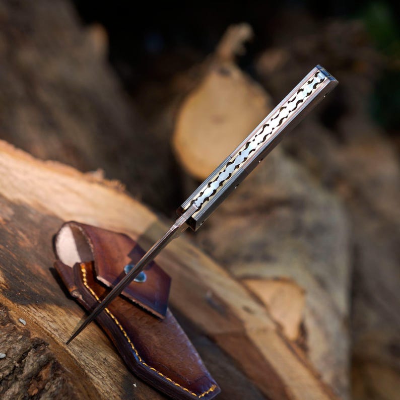 May include: A folding knife with a metallic handle featuring a decorative scalloped spine pattern. The knife is partially inserted into a brown leather sheath with visible yellow stitching and a metal rivet detail on the front flap.
