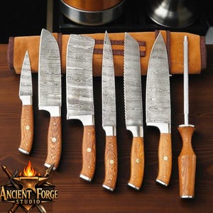 Può includere: Un set di sei coltelli da cucina e un acciaino con lame in acciaio damasco e manici in legno marrone chiaro. Ogni coltello presenta un distinto motivo ondulato sul metallo. Il set è disposto davanti a una custodia avvolgibile in pelle.