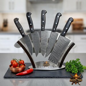 Puede incluir: Un juego de cinco cuchillos de cocina con hojas de acero damasco y mangos de madera oscura, dispuestos en un soporte de metal curvo con el logotipo de Ancient Forge Studio. Los cuchillos varían en tamaño y forma, incluyendo un cuchillo carnicero.