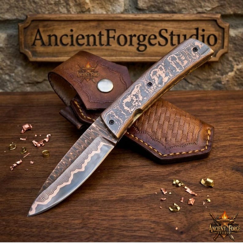 May include: A folding knife with a patterned copper and steel blade and a matching copper handle, resting on a brown leather sheath with a silver snap button. The blade features a wavy layered design, and the handle has a textured finish.