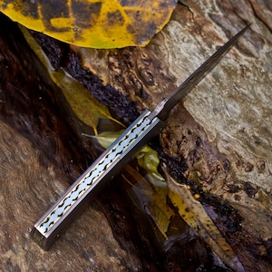 May include: A folding pocket knife with a dark metallic blade and a handle featuring a decorative, scalloped brass-colored spine inlay. The knife is positioned diagonally, showcasing the intricate metalwork along the back of the handle.