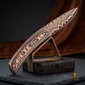 May include: A folding knife featuring a blade and handle with a distinct copper and black wavy Damascus pattern. The handle is secured with two visible screws. The knife rests on a metallic stand marked with the text ANCIENT FORGE STUDIO.