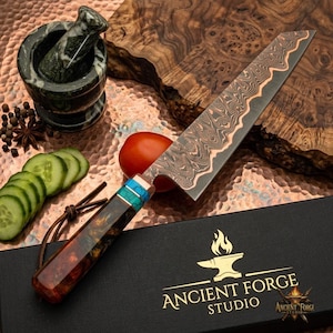Può includere: Un coltello da cucina con lama in acciaio damasco a motivi, manico in resina color ambra con accenti turchesi e una scatola di presentazione nera con il testo dorato ANCIENT FORGE STUDIO e un logo di incudine con fiamma.