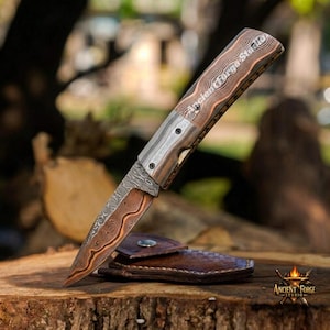 May include: A folding pocket knife with a patterned Damascus steel blade featuring copper-colored wavy lines. The handle has a layered wood grain texture with the text Ancient Forge Studio engraved on the side. A brown leather sheath lies underneath.