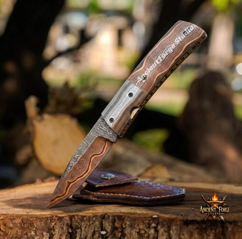 May include: A folding pocket knife with a patterned Damascus steel blade featuring copper-colored wavy lines. The handle has a layered wood grain texture with the text Ancient Forge Studio engraved on the side. A brown leather sheath lies underneath.