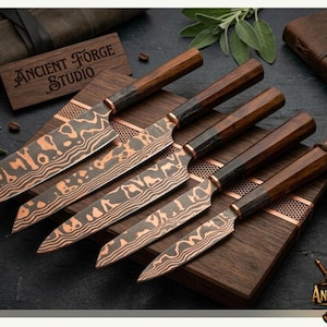 May include: A set of five kitchen knives with copper-toned Damascus patterned blades and dark wooden handles. The knives are arranged on a dark wooden block. A small wooden plaque with the text Ancient Forge Studio is visible near the knives.