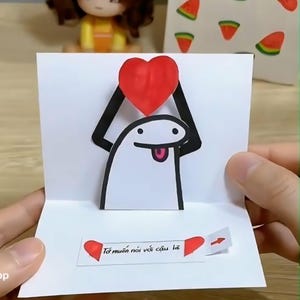 May include: A white pop-up greeting card with a cartoon figure holding a red heart. The card has black accents and Vietnamese text. A small gift bag with a watermelon pattern is visible in the background.