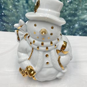 May include: A white ceramic snowman figurine with gold accents. The snowman wears a top hat, scarf, and coat, and holds a bag. The figurine has gold details on the buttons, scarf, and bag.