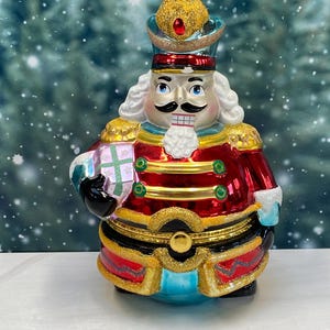 May include: A decorative Christmas ornament shaped like a nutcracker soldier. The ornament features a red jacket with gold trim, a black belt, and a gold crown. It holds a small, wrapped gift. The nutcracker has a white wig, a black mustache, and a smiling mouth.