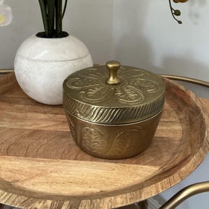 May include: A round brass container with a matching lid featuring an embossed fern-like leaf pattern. The lid has a central knob handle, and the sides of the container and lid are decorated with repeating etched botanical motifs in a metallic gold finish.